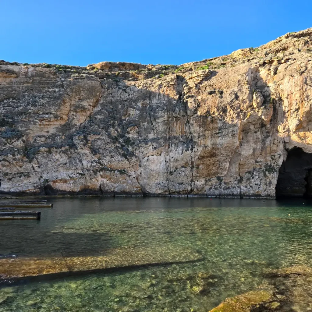 O imagine cu Vedere la Marea Interioară și stâncile din Dwejra, Gozo, Malta. Arată o lagună de apă sărată înconjurată de stânci înalte de calcar, conectată la mare printr-un tunel natural îngust.