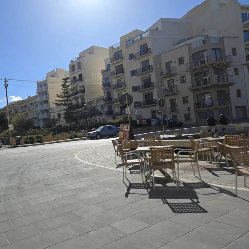 O stradă însorită din Marsalforn, Gozo, cu clădiri de apartamente, o terasă de cafenea cu scaune goale și o mașină parcată.
