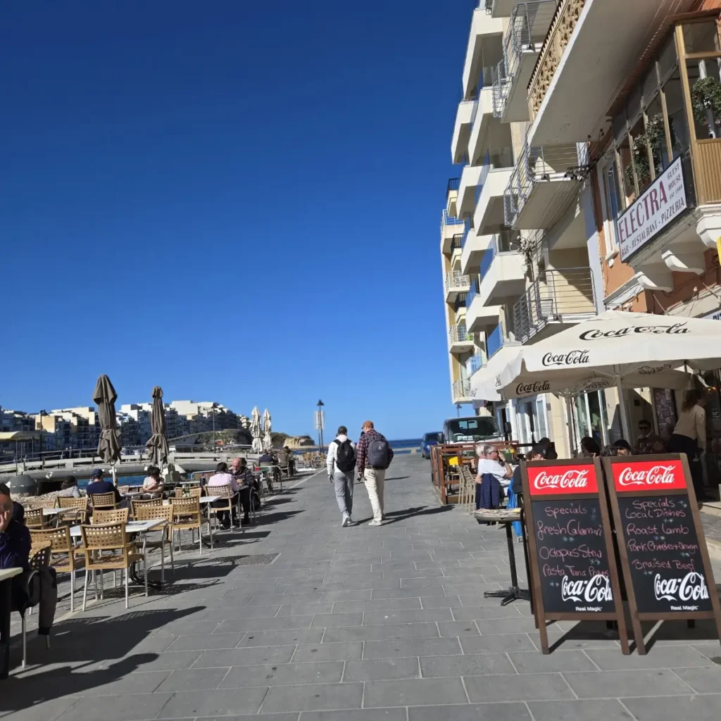 O stradă pietonală însorită, mărginită de restaurante și bărci, în Marsalforn, cu oameni așezați la mese și placi cu meniul restaurantului Electra.