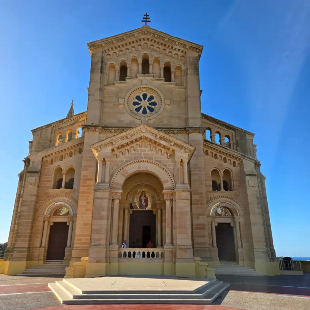 Fațada magnifică a Bazilicii Ta' Pinu din Gozo, Malta, o biserică de pelerinaj renumită, construită din piatră de nisip, sub un cer albastru senin.