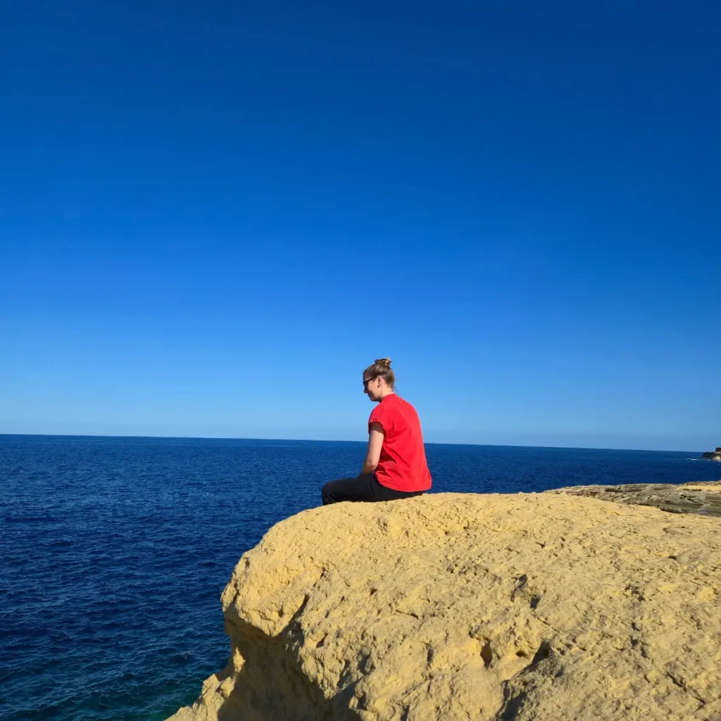 Claudia pe marginea unei stânci, privind spre marea albastră, sub un cer senin. Persoana poartă un tricou roșu.