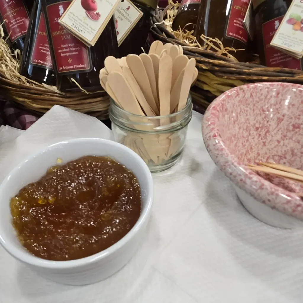 Un bol mic alb plin cu gem de rodie Savina, alături de un borcan de sticlă cu linguri de lemn și un alt bol de ceramică roz cu pete.