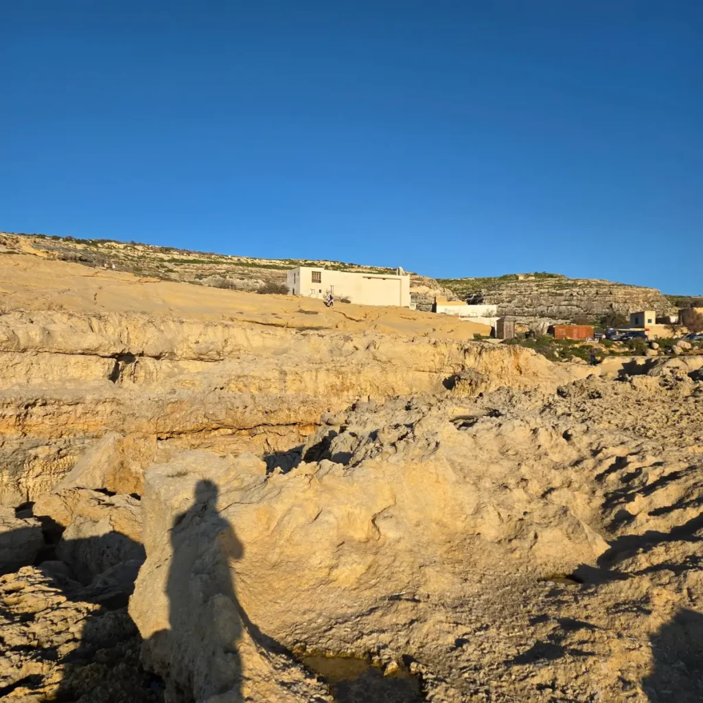O vedere panoramică a coastei stâncoase din Dwejra, Malta, sub un cer albastru senin, cu umbra unei persoane în prim-plan.