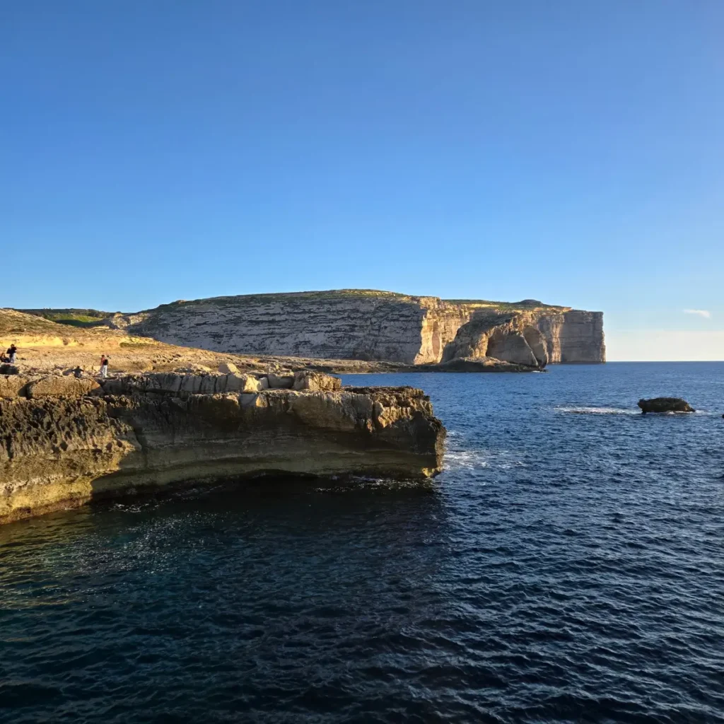 Faleză stâncoasă și mare cristalină în Golful Dwejra, Malta. Imaginea surprinde peisajul natural impresionant al coastei, cu formațiuni de rocă masive contrastând cu albastrul intens al apei.
