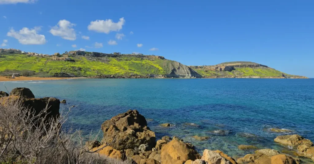 O vedere panoramică a golfului Ramla (Ramla Bay) din Gozo, Malta, cu plajă de nisip roșiatic, ape azurii cristaline și dealuri verzi luxuriante pe fundal.