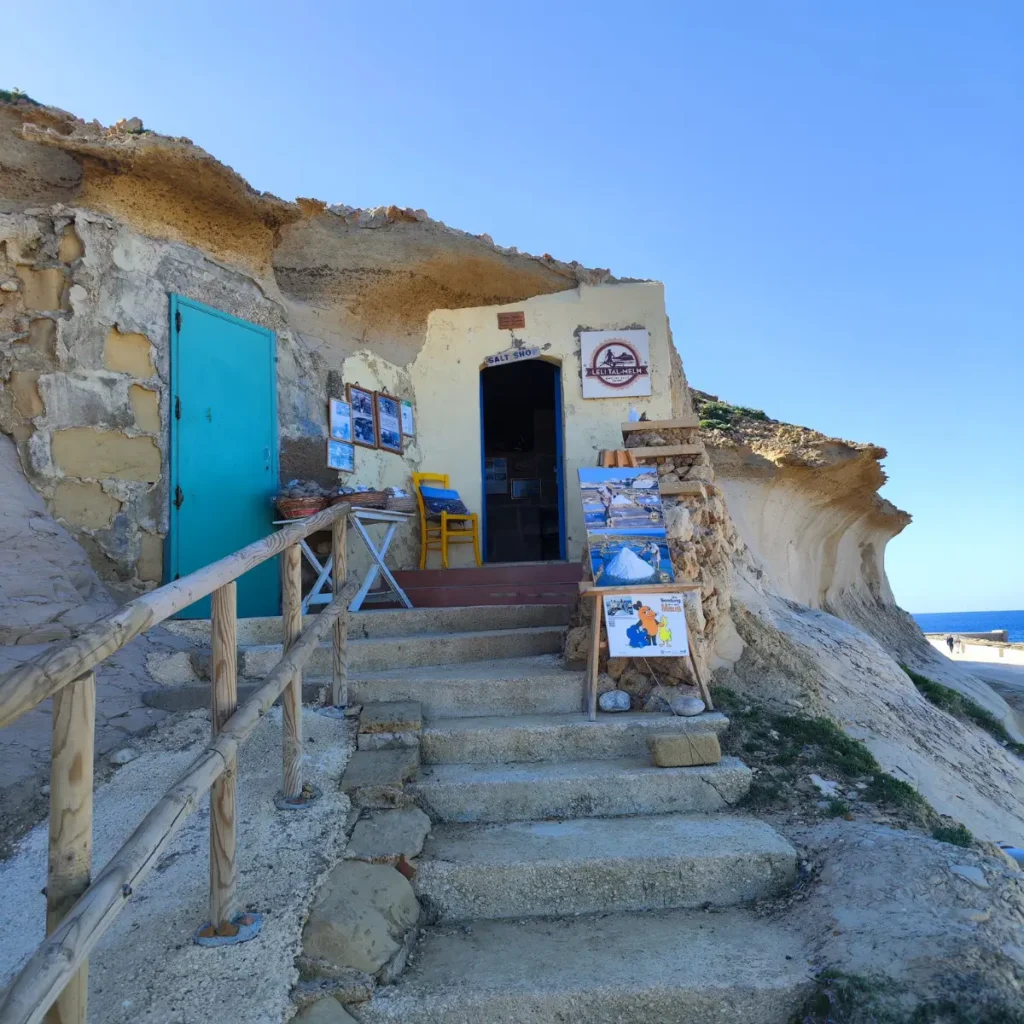 O imagine detaliată a magazinului de sare Leli Tal-Melh, sculptat direct în stâncă, cu trepte de piatră care duc la intrare și salinele Xwejni vizibile în fundal, pe coasta de nord a insulei Gozo, Malta.