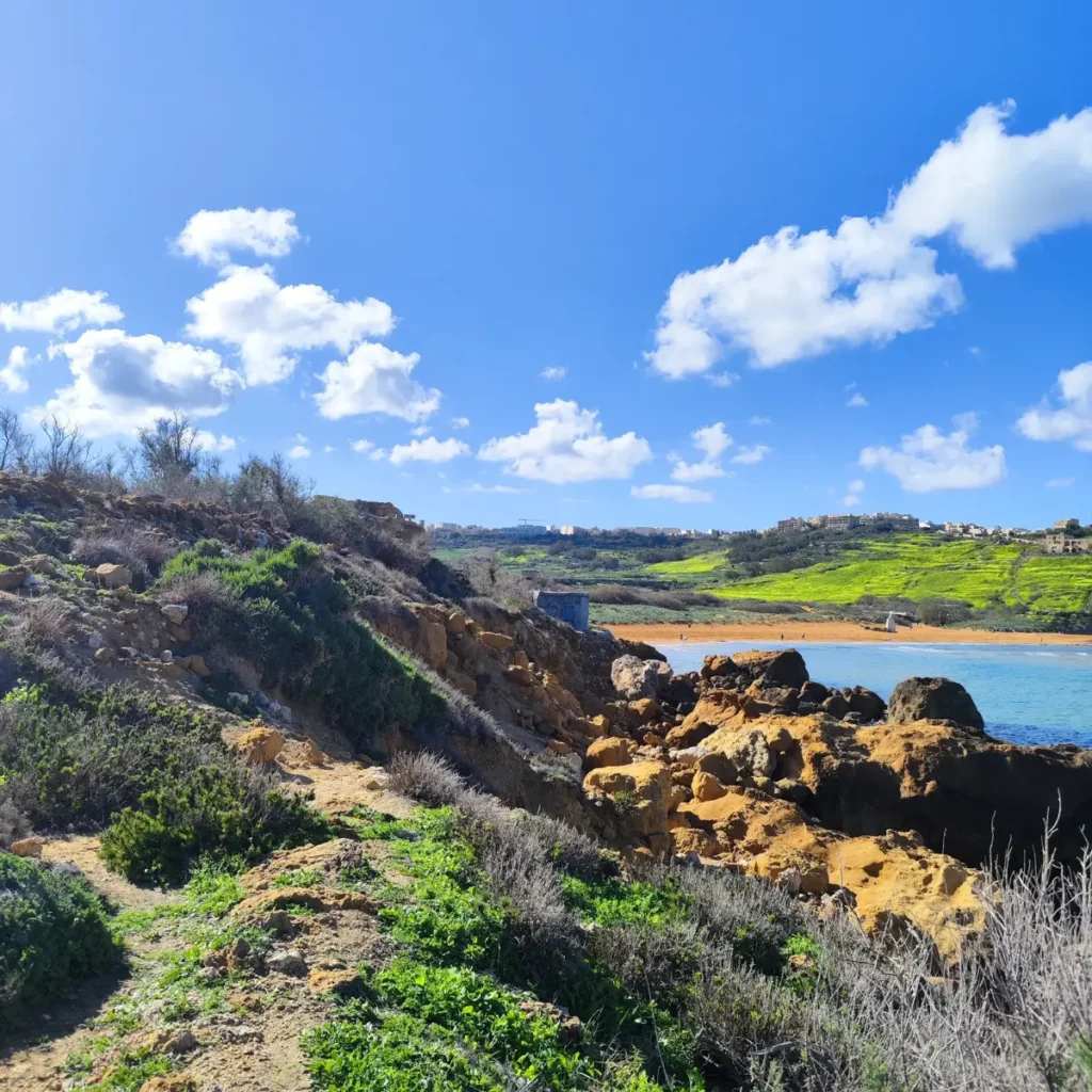 O vedere uimitoare a golfului Ramla din Gozo, cu nisipul său roșiatic-auriu întins de-a lungul coastei, atins ușor de apele turcoaz. Imaginea surprinde dealurile verzi luxuriante presărate cu flori sălbatice galbene vibrante. Golful este renumit pentru nisipul său unic de culoare roșiatică, de unde și numele său maltez, "Ramla l-Ħamra", care se traduce prin "plaja cu nisip roșu