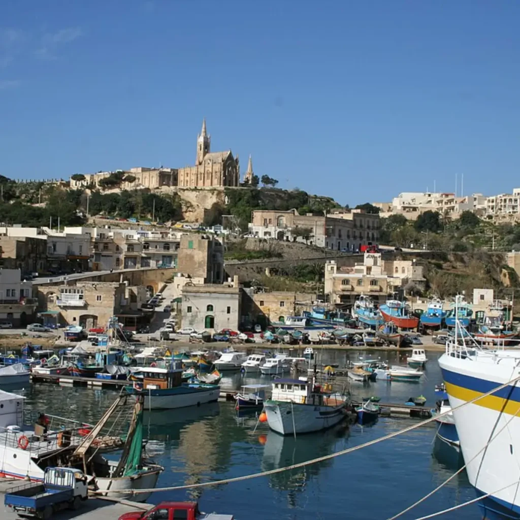 Portul Mgarr din Gozo, Malta, cu bărci de pescuit colorate și clădiri tradiționale pe un deal sub cer senin.