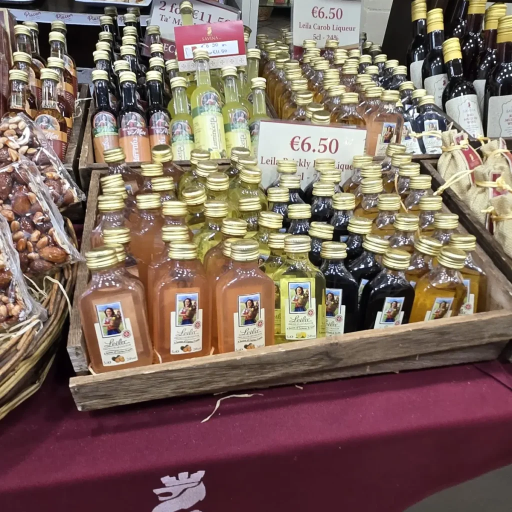 O expunere aglomerată de sticle mici de lichior, inclusiv lichior de roșcove Leila Carob Liqueur și Lumincetta, de la brandul Savina din Gozo, cu etichete de preț de 6,50 € vizibile.