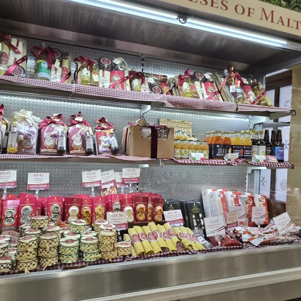 O imagine detaliată a unui stand de prezentare a produselor alimentare la Savina Artisan Centre Gozo, care prezintă o selecție variată de brânzeturi Gozo ambalate, sticle de băuturi și alte specialități locale, cu etichete de preț vizibile.