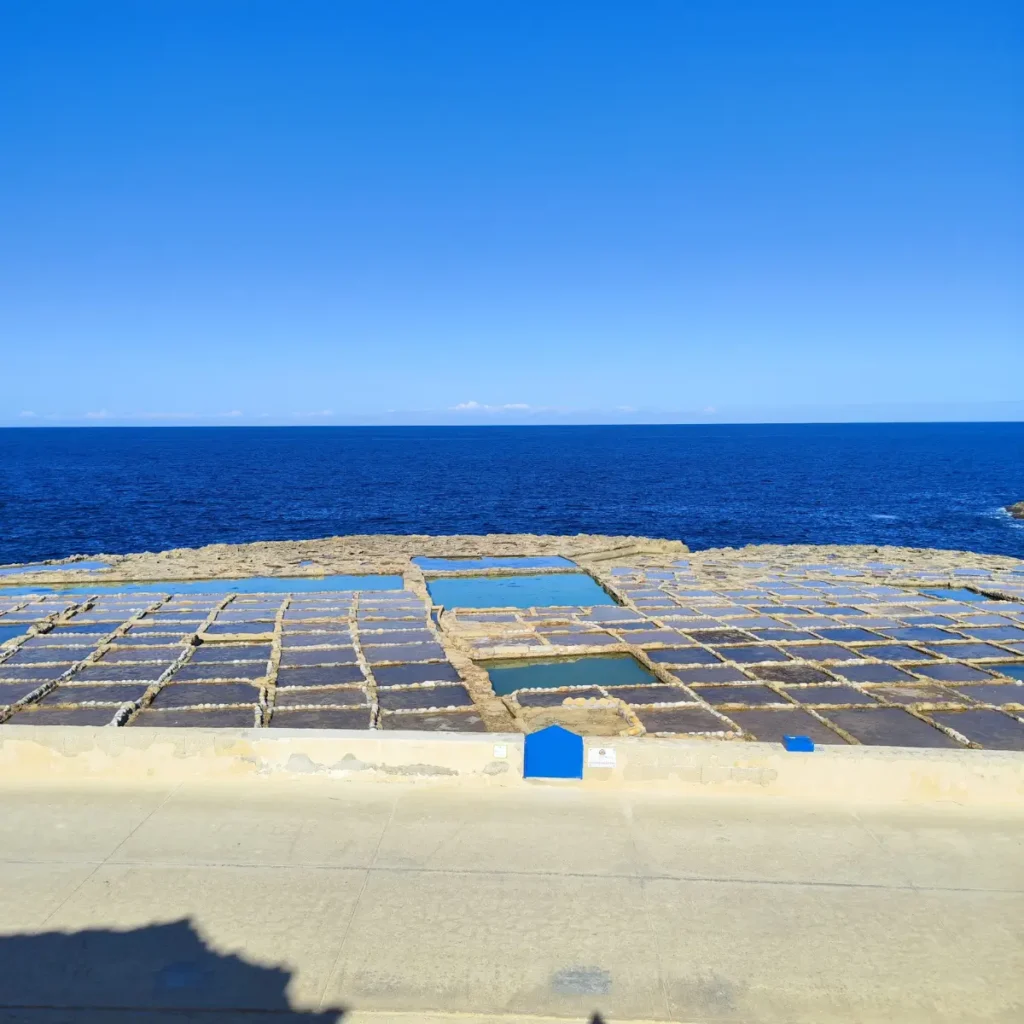 O imagine detaliată a salinelor iconice de la Xwejni, Gozo, sculptate în stânca de coastă, cu bazine geometrice care colectează apa mării pentru producția de sare, contrastând cu albastrul intens al Mării Mediterane.