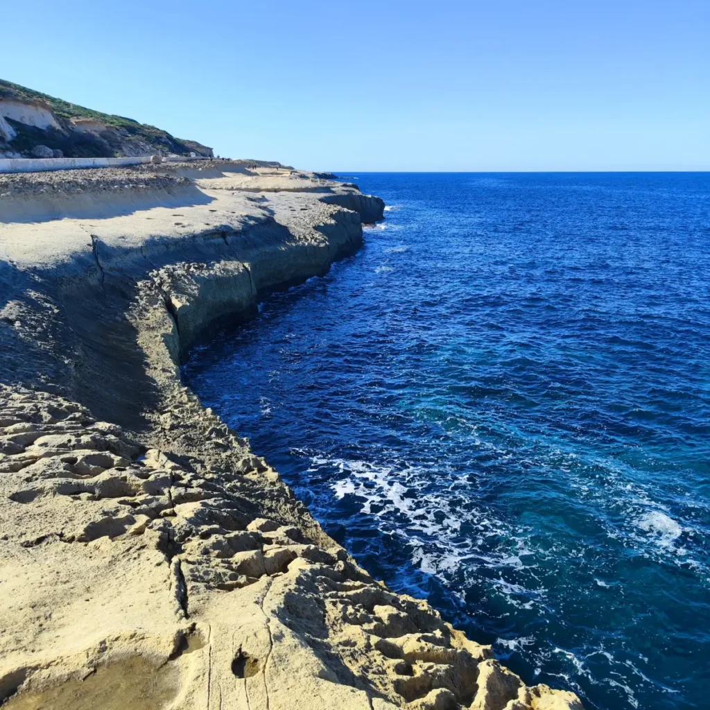 O vedere panoramică a coastelor stâncoase din Xwejni, Gozo, Malta, cu marea Mediterană de un albastru intens și cer senin. În prim-plan sunt vizibile formațiuni de stâncă erodate și posibile bazine de evaporare a sării sculptate în rocă.
