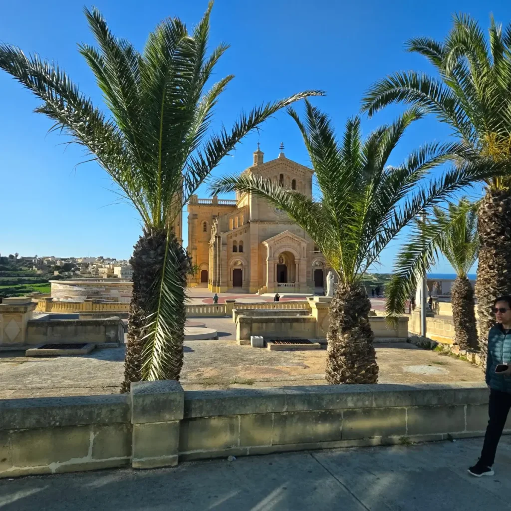O imagine panoramică luminoasă a Bazilicii Ta' Pinu din Gozo, Malta, în stil neo-romantic, cu fațada din calcar de culoarea mierii, încadrată de frunze mari de palmier pe un cer albastru senin.