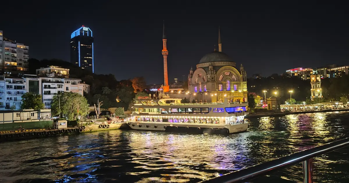 Istanbul Sub Stele: Eleganța Nocturnă a Bosforului O moschee grandioasă și un vas de croazieră iluminat pe Bosfor, Istanbul, noaptea, cu reflexiile luminilor pe apă și clădiri moderne în fundal, creând o panoramă spectaculoasă.