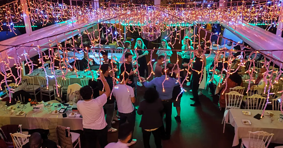 Distracție Nocturnă: Petrecere pe Croaziera Bosfor Petrecere vibrantă pe puntea unui vas de croazieră pe Bosfor, cu oaspeți dansând sub o multitudine de lumini festive și un glob disco, creând o atmosferă animată de seară.