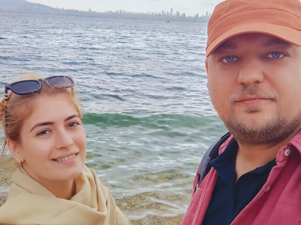 Explorând Paradisul: Noi pe Insulele Prinților Doi călători fericiți, eu și Claudia, zâmbind pe malul apei cristaline pe una dintre pitoreștile Insule ale Prinților, Istanbul, cu orașul la orizont.