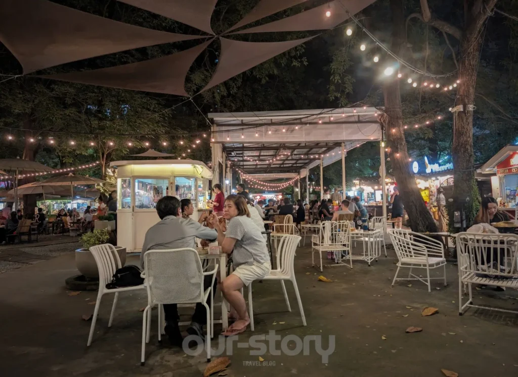 Vedere de ansamblu a pieței de mâncare The Camping Ground Bangkok, cu mese iluminate și ghirlande luminoase, noaptea