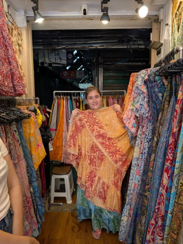 Claudia zâmbitoare ține o eșarfă colorată, cu modele roșii și aurii, într-un stand aglomerat cu haine la Piața de weekend Chatuchak din Bangkok, Thailanda.