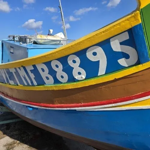 Barca colorata luzzu detaliu in Marsaxlokk, Malta, o imagine care surprinde un detaliu vibrant al unei bărci tradiționale malteze, cunoscută sub numele de luzzu, evidențiind culorile vii și designul distinctiv al acestei bărci în portul pitoresc al satului de pescari Marsaxlokk, cu clădirile colorate și cerul senin în fundal.