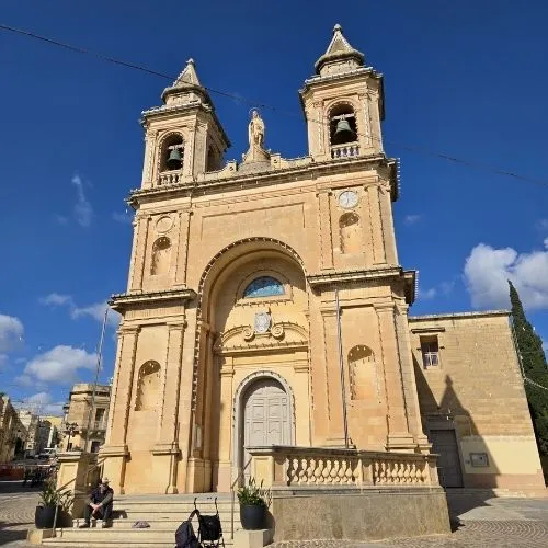 Biserica Parohiala Nostra Signora di Pompei, Marsaxlokk, Malta, o imagine care surprinde o scenă pitorească în care biserica este înconjurată de clădirile colorate ale satului, cu bărcile tradiționale malteze, cunoscute sub numele de luzzu, ancorate în portul pitoresc al satului de pescari Marsaxlokk, cu clădirile colorate și cerul senin în fundal.