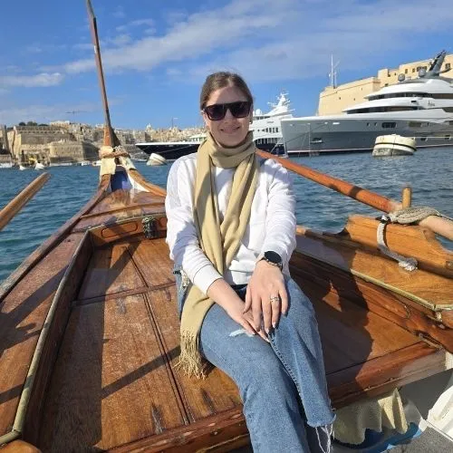 Claudia pe barca tradițională dghajsa, Malta, o imagine care surprinde un moment de plimbare pe apele liniștite ale portului, evidențiind atmosfera sa relaxantă și farmecul său autentic, cu bărci colorate care navighează în fundal.