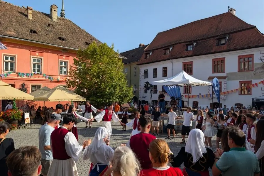 Bogdan și Claudia pe zidul cetății Sighișoara cu marea de acoperișuri medievale din țiglă roșie și dealurile Transilvaniei în fundal — Drumul Panoramic la apus