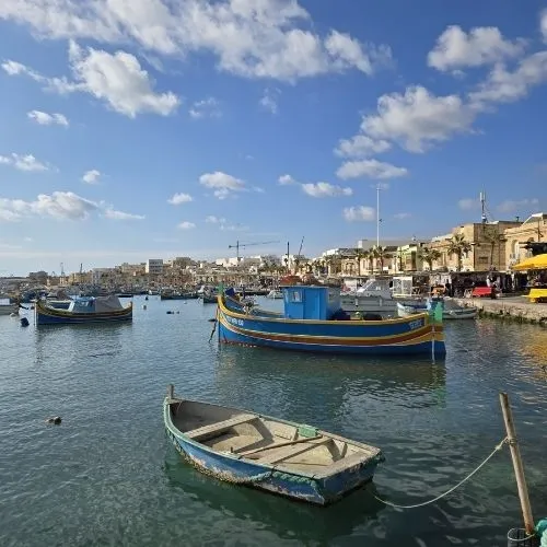 Golful Marsaxlokk și bărcute tradiționale luzzu, Malta, o imagine care surprinde o scenă pitorească în care golful Marsaxlokk este înconjurat de bărcute tradiționale luzzu, cu clădirile colorate și cerul senin în fundal.