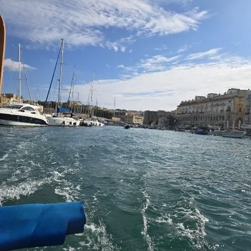 In Marele Port cu barca traditionala dghajsa, Malta, o imagine care surprinde o scenă pitorească în care o barcă tradițională malteză, cunoscută sub numele de dghajsa, navighează pe apele liniștite ale Marelui Port din Malta, cu clădirile istorice și cerul senin în fundal.