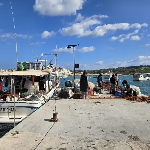 Pescari gata de a iesi in larg, Marsaxlokk, Malta, o imagine care surprinde o scenă pitorească în care pescarii sunt gata să plece în larg, cu bărcile tradiționale malteze, cunoscute sub numele de luzzu, ancorate în portul pitoresc al satului de pescari Marsaxlokk, cu clădirile colorate și cerul senin în fundal.