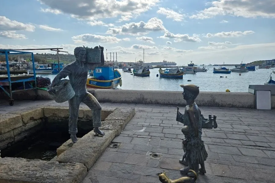 Statuete cu pescari in Marsaxlokk, Malta, o imagine care surprinde o scenă pitorească în care statuetele reprezintă pescari într-o atmosferă pitorescă, cu bărcile tradiționale malteze, cunoscute sub numele de luzzu, ancorate în portul pitoresc al satului de pescari Marsaxlokk, cu clădirile colorate și cerul senin în fundal.
