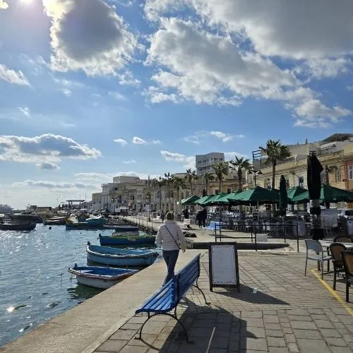 Terase în satul pescăresc Marsaxlokk din Malta, o imagine care surprinde o scenă pitorească în care terasele se aliniază de-a lungul malului portului din Marsaxlokk, cu bărcile tradiționale malteze, cunoscute sub numele de luzzu, ancorate în apele liniștite, și clădirile colorate ale satului în fundal, creând o atmosferă relaxantă și autentică.