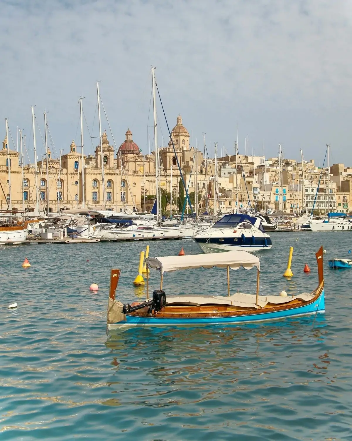 Dghajsa, barca tradițională malteză, o imagine care surprinde o barcă tradițională dghajsa în apele liniștite ale portului din Malta, evidențiind designul său distinctiv și farmecul său autentic, cu clădirile istorice și cerul senin în fundal, creând o atmosferă relaxantă și pitorească.