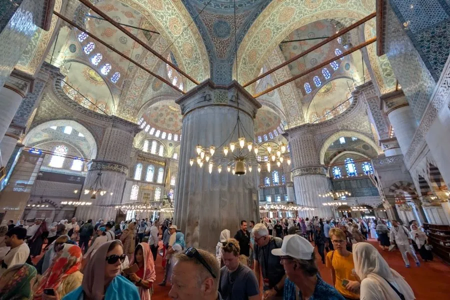 Turiști în interiorul Moscheii Albastre din Istanbul — faianță albastră Iznik și lumina naturală filtrată prin vitralii